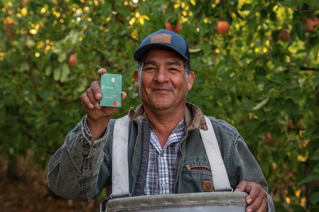 A smiling worker holding a Ganaz payroll card in an orchard